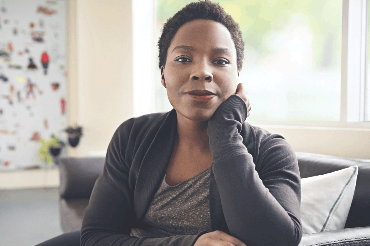 A Black Woman sitting on a sofa stares confidentally at the viewer with a half smile on her face and her head resting on her