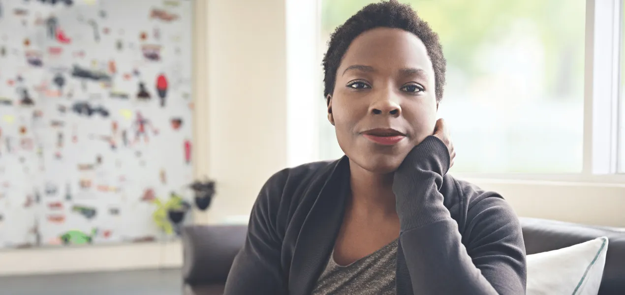 A Black Woman sitting on a sofa stares confidentally at the viewer with a half smile on her face and her head resting on her hand.