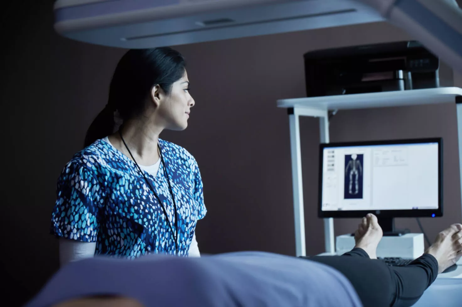 Healthcare professional looking at scan on monitor screen with patient lying on bed